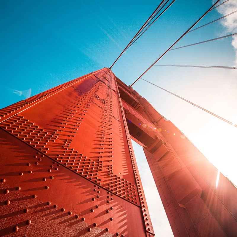 A vertical view of a bridge support with a sun burst and blue sky.